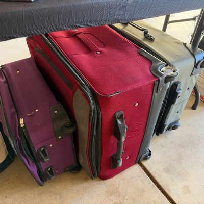 Selection of modern rolling softside suitcases in purple, red, and olive green, shown stored under a table alongside a canvas tote bag.