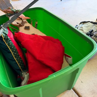 Large green plastic storage tub filled with Christmas decorations, including a festive throw pillow, red fabric tree skirt, and a wooden...