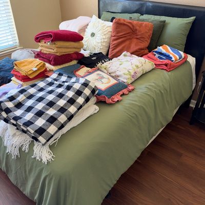 A bed displayed with stacks of colorful bath towels, throw pillows, and folded blankets, including a black-and-white buffalo plaid throw.