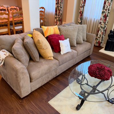 Contemporary tan microfiber three-cushion sofa styled with various throw pillows, paired with an oval glass-top coffee table on a wrought...