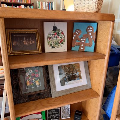 Sale Photo Thumbnail #106: A light oak two-shelf bookcase filled with framed art prints and decorative canvas paintings.