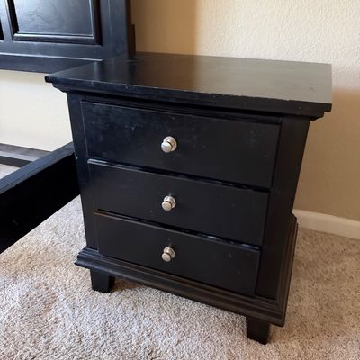 Black wooden nightstand featuring three drawers with brushed silver knobs, displaying light wear on the finish.