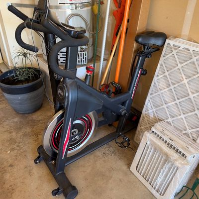 Sale Photo Thumbnail #112: Black stationary exercise bike with a weighted front flywheel, stored in a garage alongside home air filters.
