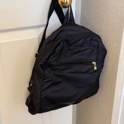 Black nylon mini backpack hanging from a doorknob, featuring multiple zippered compartments and adjustable shoulder straps.