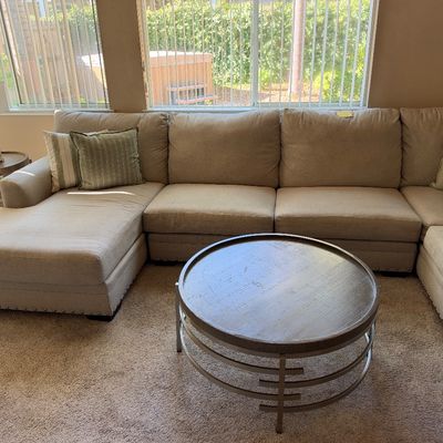 Large beige U-shaped sectional sofa with nailhead trim and accent pillows, paired with a modern round wood and metal coffee table.