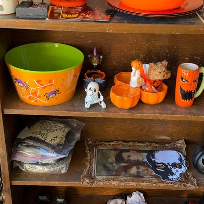 Sale Photo Thumbnail #140: A wooden shelf filled with various Halloween-themed decorations, including an orange spiderweb mixing bowl, jack-o'-lantern mugs, pumpkin-shaped bowls, small figurines, and a framed skull mask.