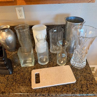 Assorted collection of glass, ceramic, and metal vases alongside a small rectangular cutting board arranged on a speckled countertop.