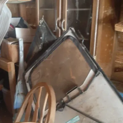 Cluttered storage area featuring a vintage wooden display cabinet with glass doors, a wooden chair, and automotive parts, including a...