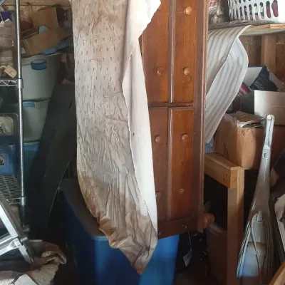 Tall vintage wooden dresser partially covered with fabric, stored in a shed alongside plastic totes, boxes, and miscellaneous household...