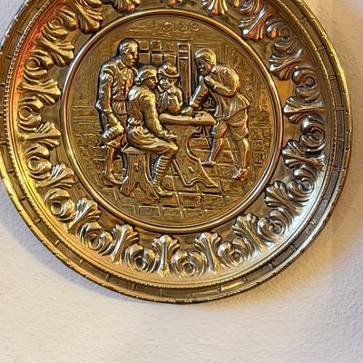 Vintage embossed brass wall plaque depicting a pub scene with figures seated at a table, featuring an ornate scrollwork border.