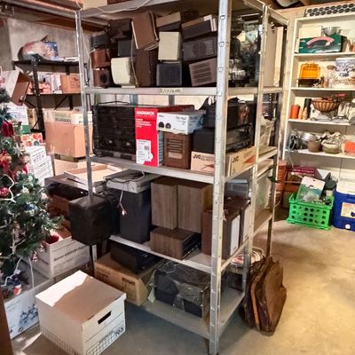 Metal shelving unit filled with vintage electronics, including radios, stereo receivers, speakers, and boxed audio equipment in a...
