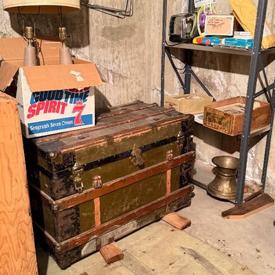 Antique flat-top steamer trunk with wood bands and brass hardware, shown alongside a metal shelving unit and a vintage table lamp in a...
