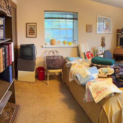 Bedroom filled with household goods, including a tall wooden bookshelf, vintage quilts displayed on a bed, desks, and electronics.
