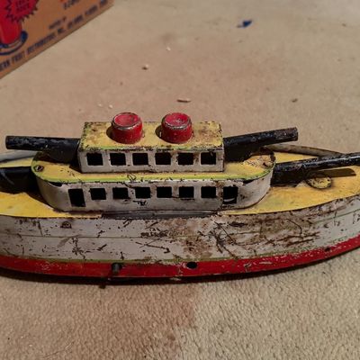 Vintage tin litho toy battleship featuring red, yellow, and white painted details, showing significant paint wear and surface patina from...