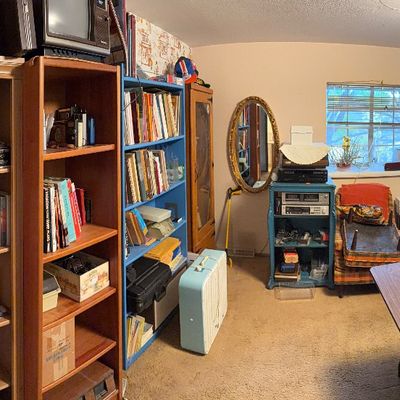 Home office and study filled with wooden bookcases, books, vintage electronics, a portable fan, and an oval wall mirror.