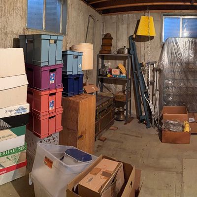 Unfinished basement storage area filled with stacked cardboard boxes, colorful plastic crates, metal shelving units, crutches, and...