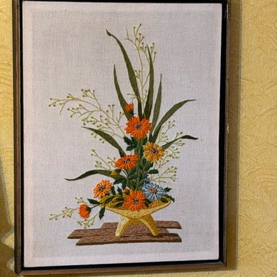 Vintage framed crewel embroidery depicting an arrangement of orange and yellow flowers in a tripod vase, housed in a simple dark wood frame.