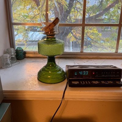 Vintage green textured glass oil lamp with a clear glass chimney, sitting on a windowsill next to a digital alarm clock radio.
