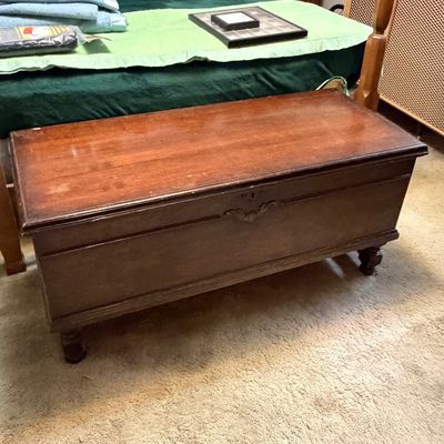 Vintage dark wood cedar chest with carved trim detail and turned feet, positioned at the end of a bed.
