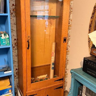 Tall wooden gun cabinet featuring a glass-front display door, bottom storage cabinet, and black wrought iron hardware.