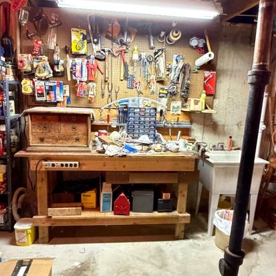 Basement workshop setup featuring a heavy-duty wooden workbench, overhead fluorescent light, pegboard wall with hanging hand tools and...
