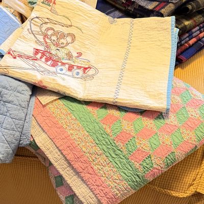 Collection of vintage linens and textiles, featuring hand-embroidered baby quilts, colorful patchwork coverlets, and plaid wool blankets.
