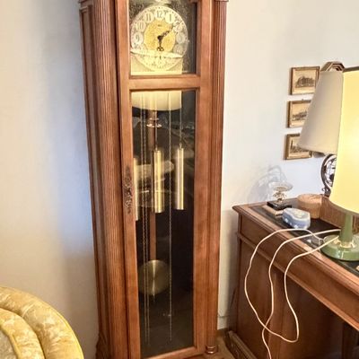Traditional wooden grandfather clock featuring a bonnet pediment, fluted columns, ornate brass dial, and visible brass weights and pendulum.