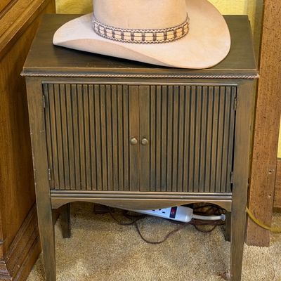 Beige cowboy hat with a braided band resting atop a small dark wood cabinet featuring tambour-style double doors.
