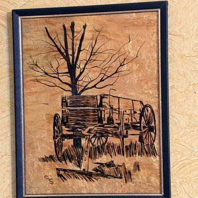 Framed pyrography or ink illustration on wood depicting a rustic wooden wagon beside a leafless tree, signed CS in the lower left corner.