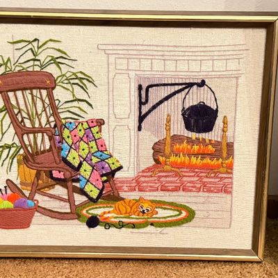 Framed vintage crewel embroidery piece depicting a cozy fireplace scene with a rocking chair, quilt, sleeping cat, and knitting basket.