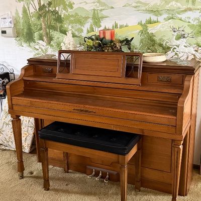 Vintage spinet upright piano in a light wood finish, complete with a matching bench featuring a black upholstered cushioned top.