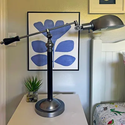 Sale Photo Thumbnail #215: Modern industrial-style adjustable boom desk lamp with a brushed nickel metal shade and black column base, positioned on a white side table.