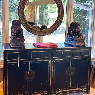 Sale Photo Thumbnail #229: Black distressed Asian-style sideboard cabinet with brass hardware, topped by a pair of carved foo dog statues and a round ornate gold mirror.