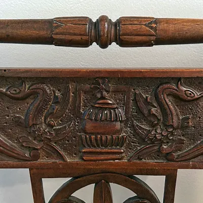 Sale Photo Thumbnail #200: Detail view of an antique carved wooden chair featuring a textured crest rail depicting stylized birds, urn motifs, and a turned spindle top.