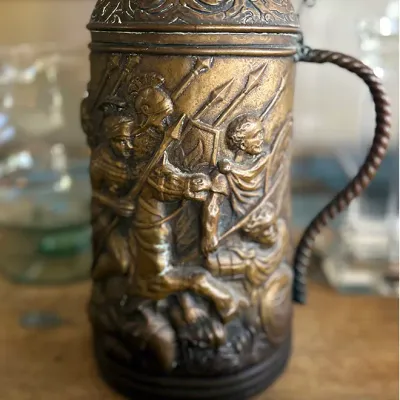 Sale Photo Thumbnail #137: Ornate antique brass lidded stein featuring relief-carved scenes of soldiers in battle with a twisted rope-style handle.