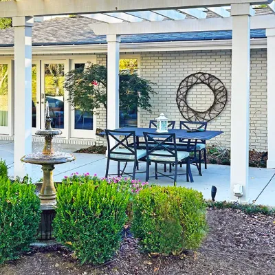 Sale Photo Thumbnail #217: Outdoor patio dining set situated under a white pergola, featuring a rectangular blue table and four cross-back chairs with light blue cushions, alongside a three-tiered concrete water fountain.