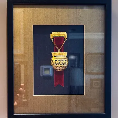 Sale Photo Thumbnail #103: Framed vintage German gold-toned commemorative ribbon badge featuring ornate filigree detailing and a crimson velvet ribbon, presented in a black shadowbox frame with a double mat.