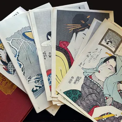 Sale Photo Thumbnail #113: Collection of Japanese ukiyo-e style woodblock prints and art reproductions spread over a red cloth presentation folder.