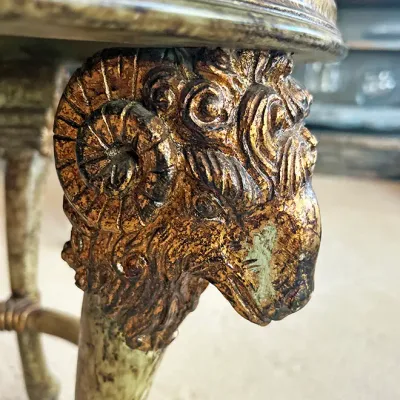 Sale Photo Thumbnail #112: Detail of an ornate gilt and painted accent table featuring a carved ram's head design at the top of the leg.