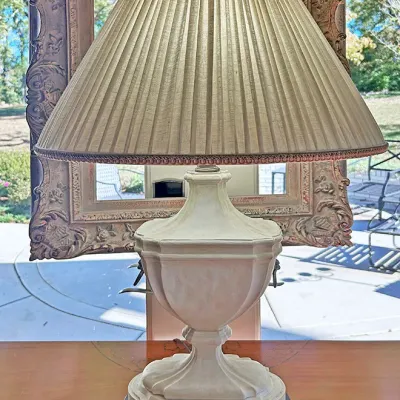Sale Photo Thumbnail #78: Neoclassical style off-white urn-form table lamp with a pleated beige lampshade, displayed in front of an ornate gilt frame.