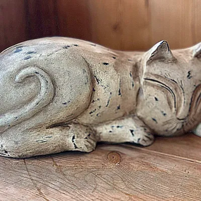 Sale Photo Thumbnail #121: Rustic carved ceramic or stone sleeping cat figurine with a weathered distressed finish, displayed on a wooden shelf.