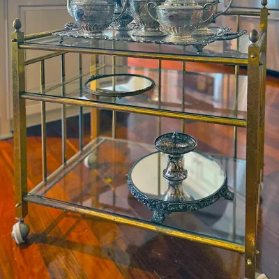 Sale Photo Thumbnail #54: Mid-century brass and glass three-tier rolling bar cart displaying an ornate silver-plated tea set and accessories.