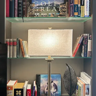 Sale Photo Thumbnail #37: Contemporary brushed nickel table lamp with a rectangular fabric shade, displayed on a bookshelf flanked by various hardcovers, bookends, and decorative sculptures.