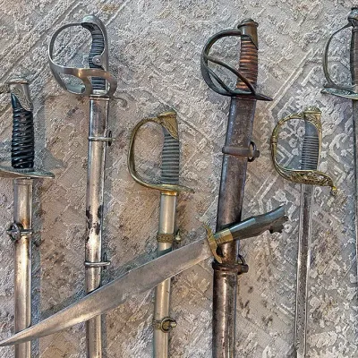 Sale Photo Thumbnail #298: Collection of antique and vintage military swords and sabers with metal scabbards, alongside a short hunting dagger, arranged on a patterned rug.