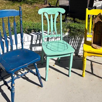Sale Photo Thumbnail #247: Trio of vintage wooden dining chairs repainted in distressed blue, mint green, and bright yellow finishes, featuring a wire lampshade frame and a rustic wooden birdhouse sitting on top.