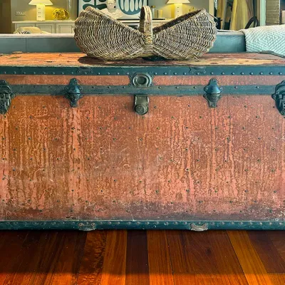 Sale Photo Thumbnail #73: Antique steamer trunk featuring a distressed reddish finish, dark metal trim, riveted details, and heavy latches, topped with a woven wicker basket.