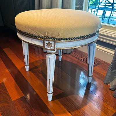Sale Photo Thumbnail #75: French Neoclassical-style round vanity stool featuring painted white and gold-finished fluted legs, nailhead trim, and beige textured upholstery.
