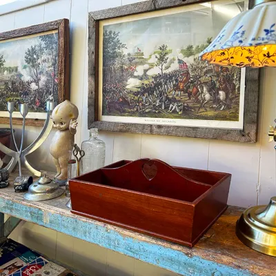 Sale Photo Thumbnail #284: Collection of vintage decor displayed on a painted rustic bench, including framed Civil War battle prints, a brass gooseneck desk lamp with a blue-and-white shade, a wooden caddy, and various small figurines.