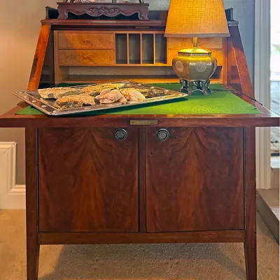 Sale Photo Thumbnail #148: Vintage drop-front mahogany secretary desk featuring a green felt writing surface, interior cubbies, and lower cabinet doors. Displayed on top is a small brass table lamp alongside decorative trays and collectibles.