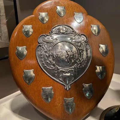 Sale Photo Thumbnail #60: Vintage oak trophy shield featuring a central repoussé silver-tone medallion and smaller engraved winner plaques dating to the 1920s.